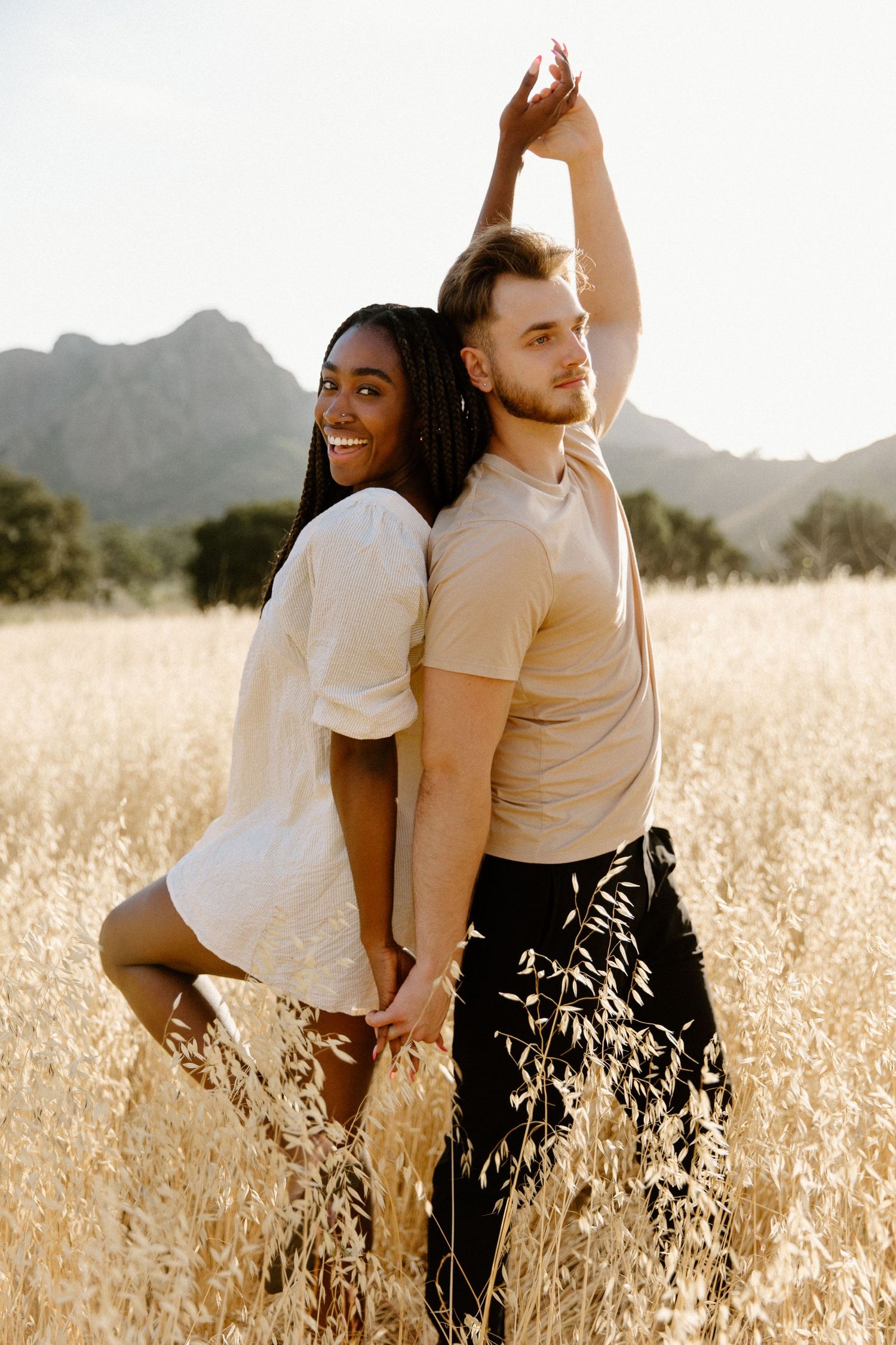 Couple stands back-to-back in a golden wheat field with mountain landscape at sunset.