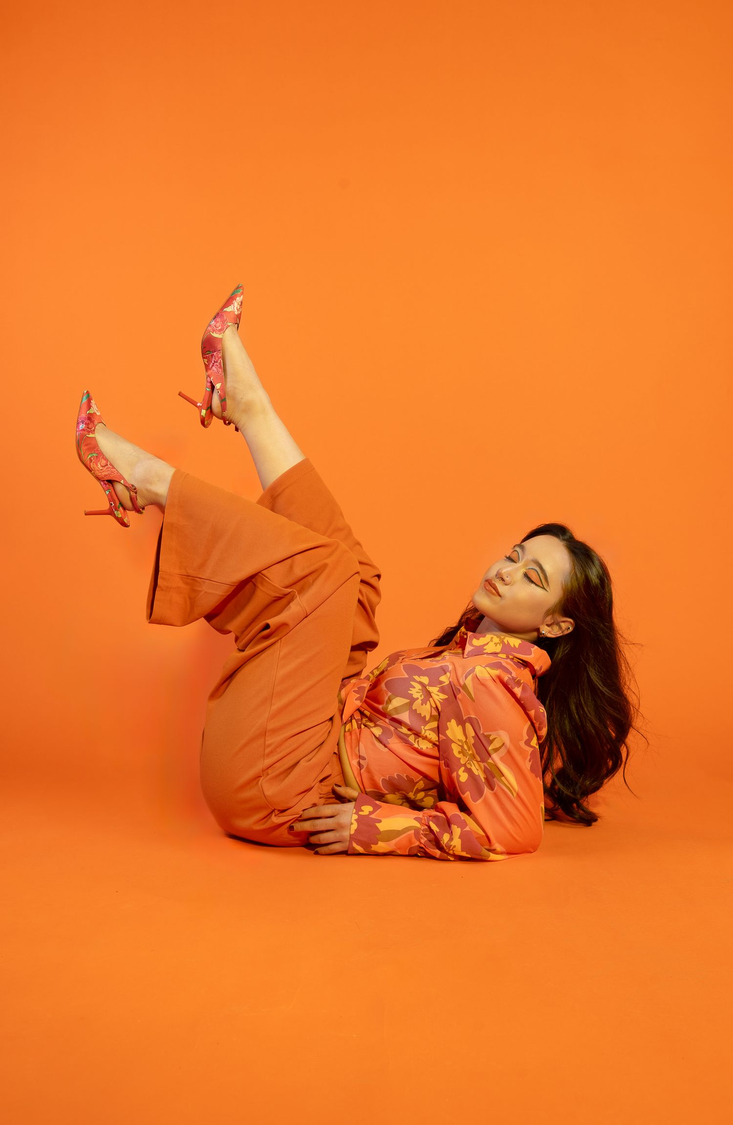 Girl posing in all orange and orange backdrop, legs in the air with eyes closed