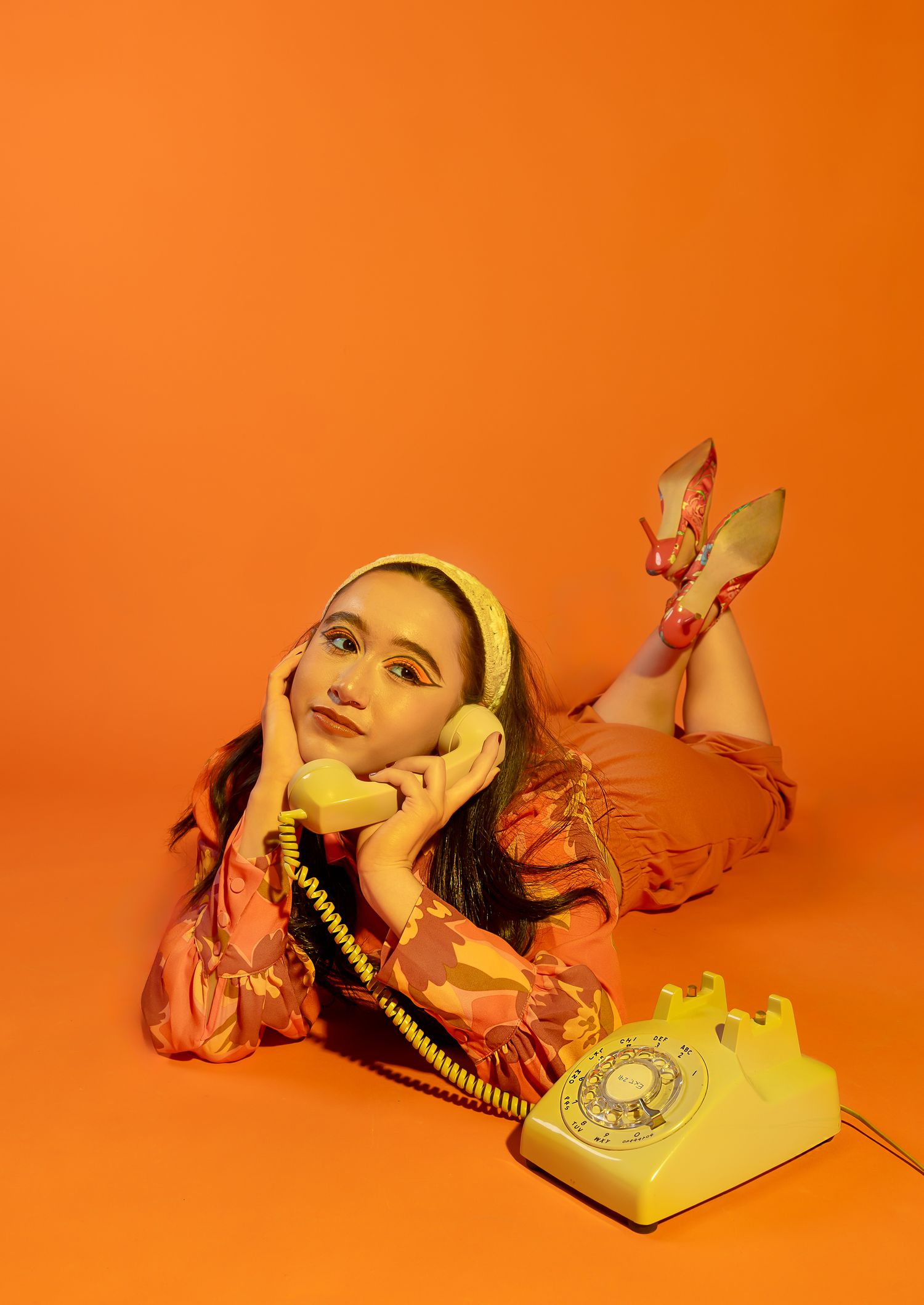 Girl posing in all orange and orange backdrop, legs crossed behind her laying on her stomach with an old telephone to her ear