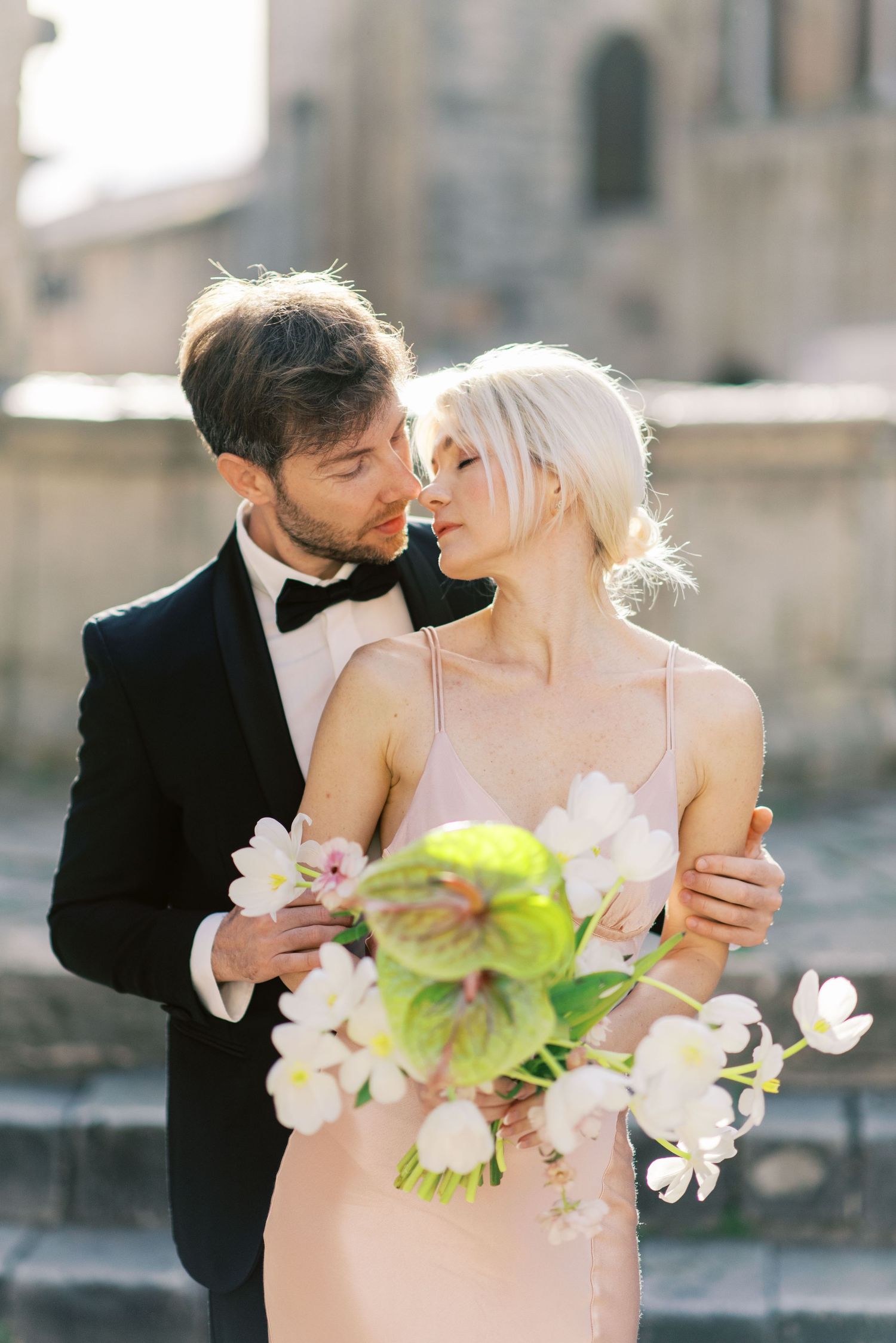 Romantic wedding couple embraces outdoors holding delicate pink and green bouquet in elegant formal attire.