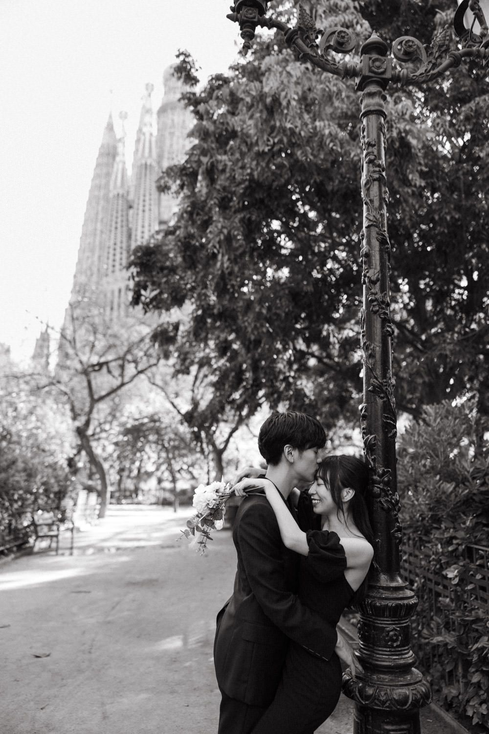 A couple shares an intimate moment by a lamppost near the Sagrada Familia in black and white.