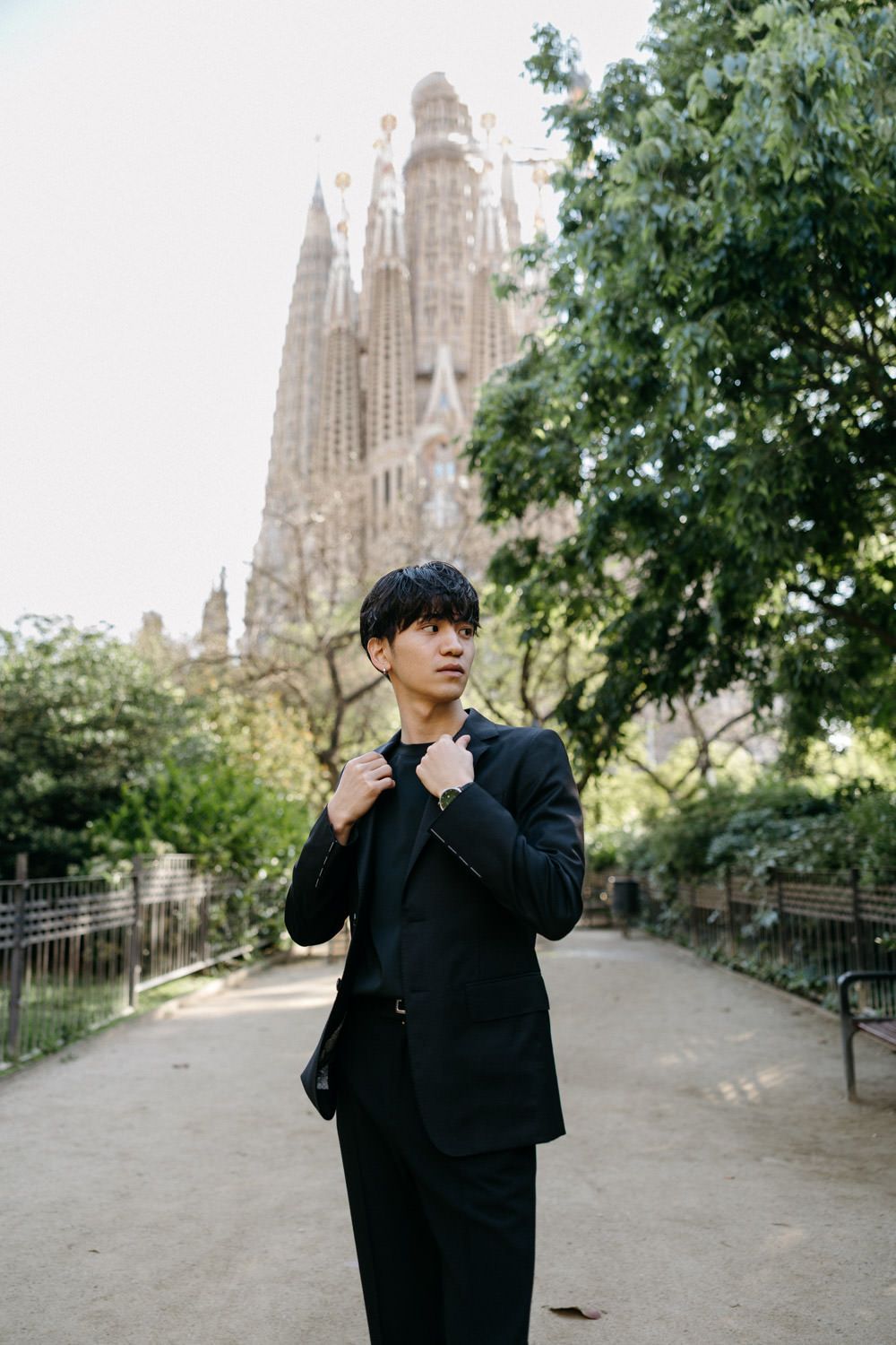 A person in a black suit poses on a path with the Sagrada Familia towers in the background.