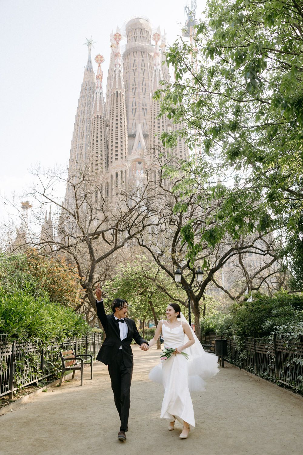 A couple in wedding attire walks along a tree-lined path with Sagrada Familia in the background.