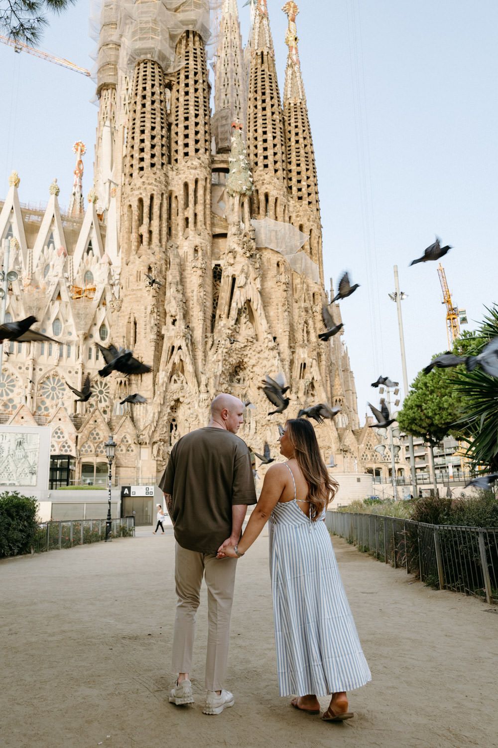 Couple admires the Sagrada Familia cathedral while pigeons fly nearby.