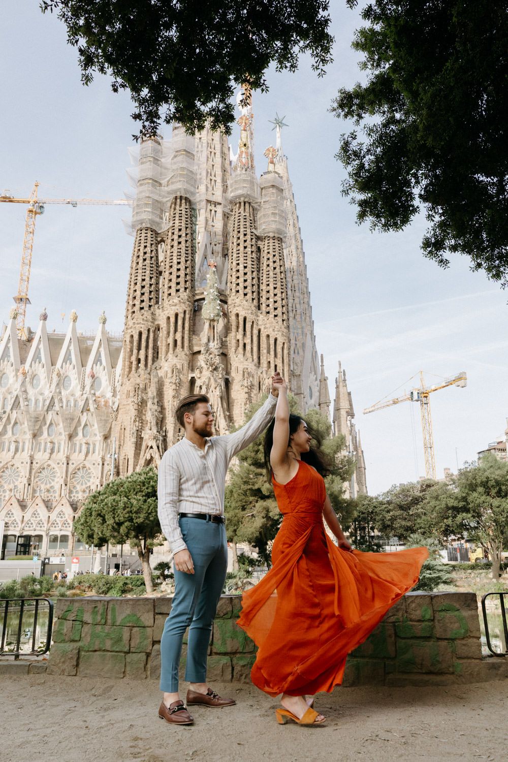 Woman in orange dress twirls dramatically with Sagrada Familia cathedral backdrop.