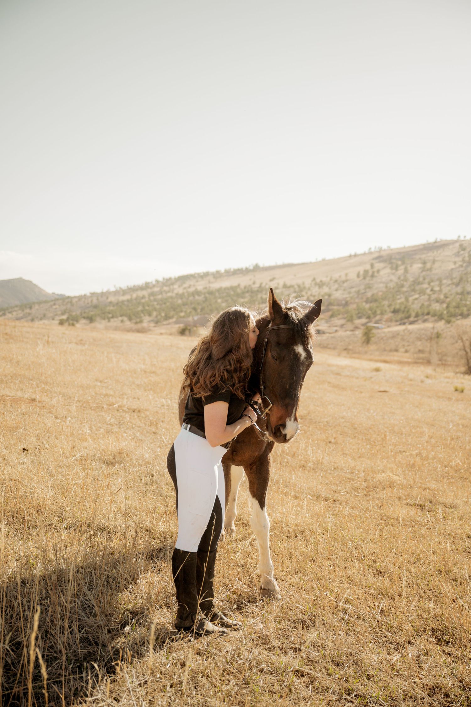 A horse and rider share an intimate moment in a golden field with mountains in the background at sunset.