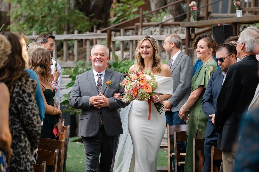 A wedding ceremony procession in an outdoor garden venue with guests gathered along the sides of the aisle.