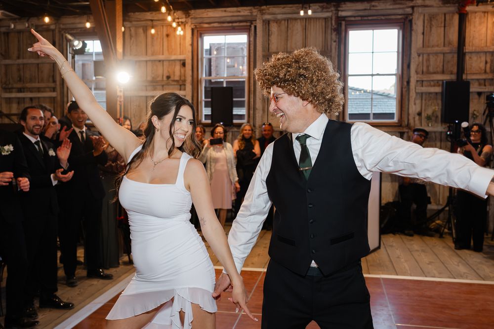 Bride and groom share joyful first dance in rustic barn wedding venue with guests celebrating in background.
