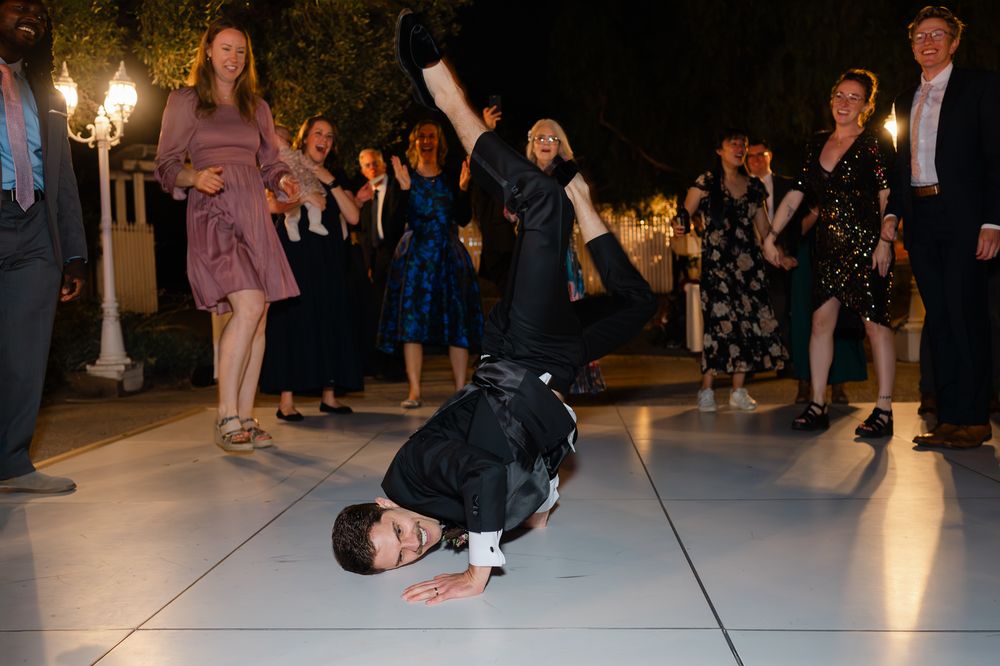 Guest breakdancing on white dance floor at wedding reception, crowd watching and cheering outdoors at night.