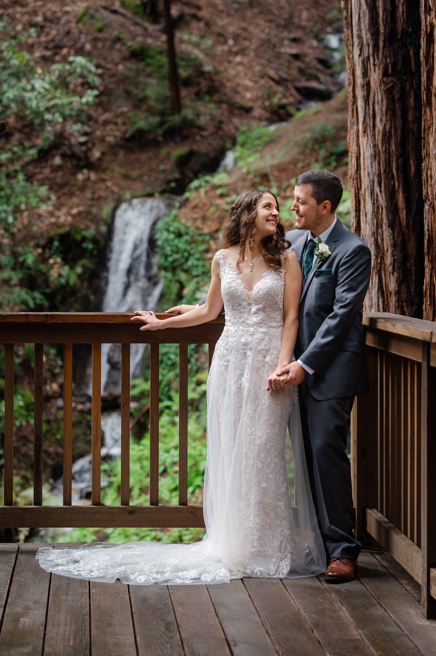 Bride and groom holding hands on a wooden deck with a waterfall and lush greenery in the background.