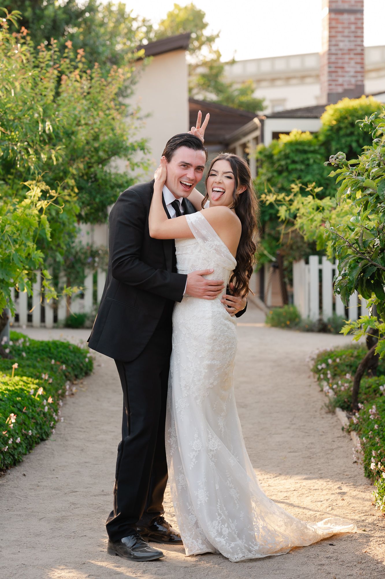 Joyful bride and groom embracing on a garden path, groom making bunny ears, both laughing in wedding attire.