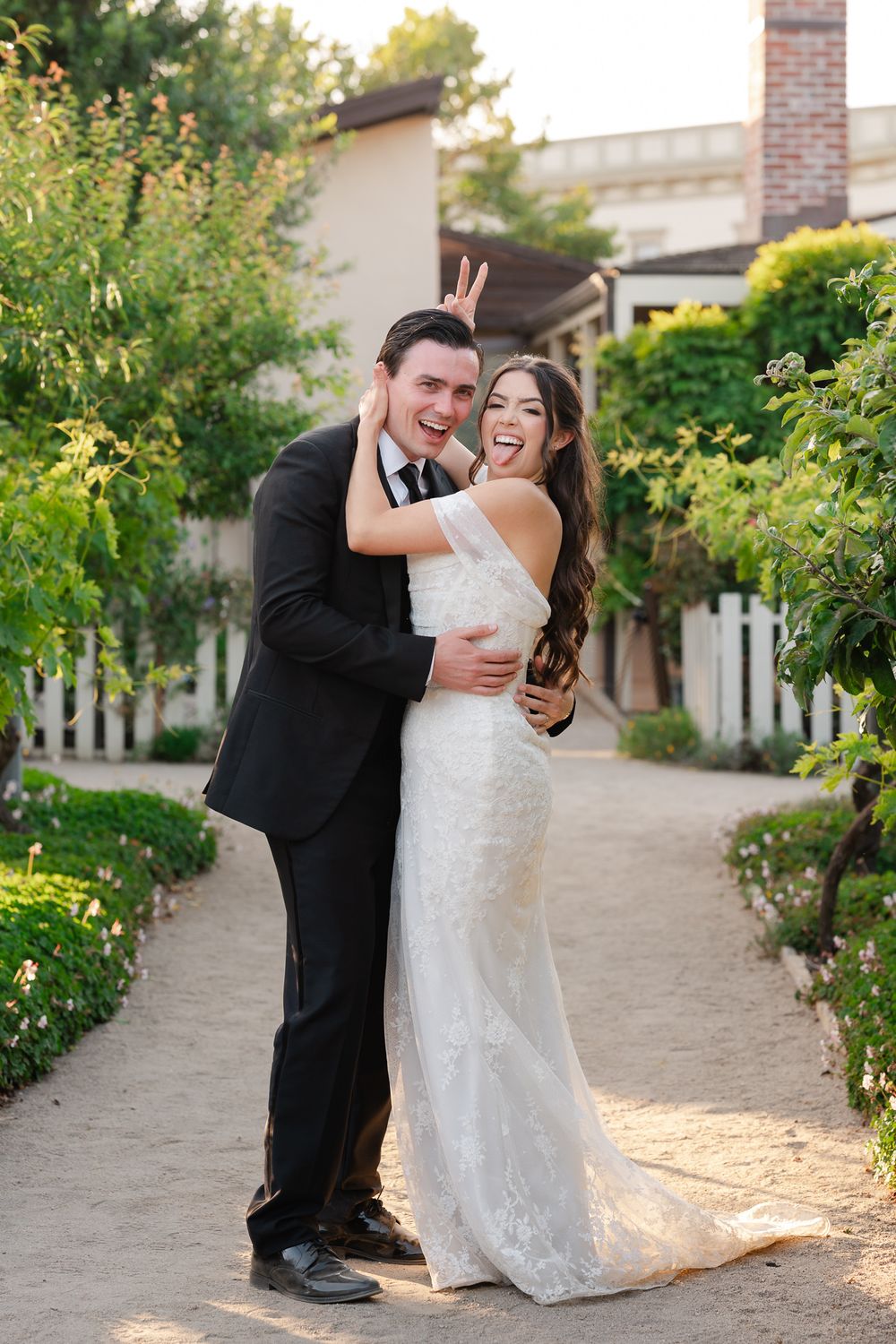 Joyful bride and groom embracing on a garden path, groom making bunny ears, both laughing in wedding attire.