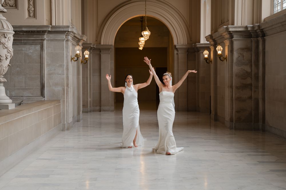 Two brides in white gowns celebrate joyfully in an elegant marble hall, tossing a bouquet.