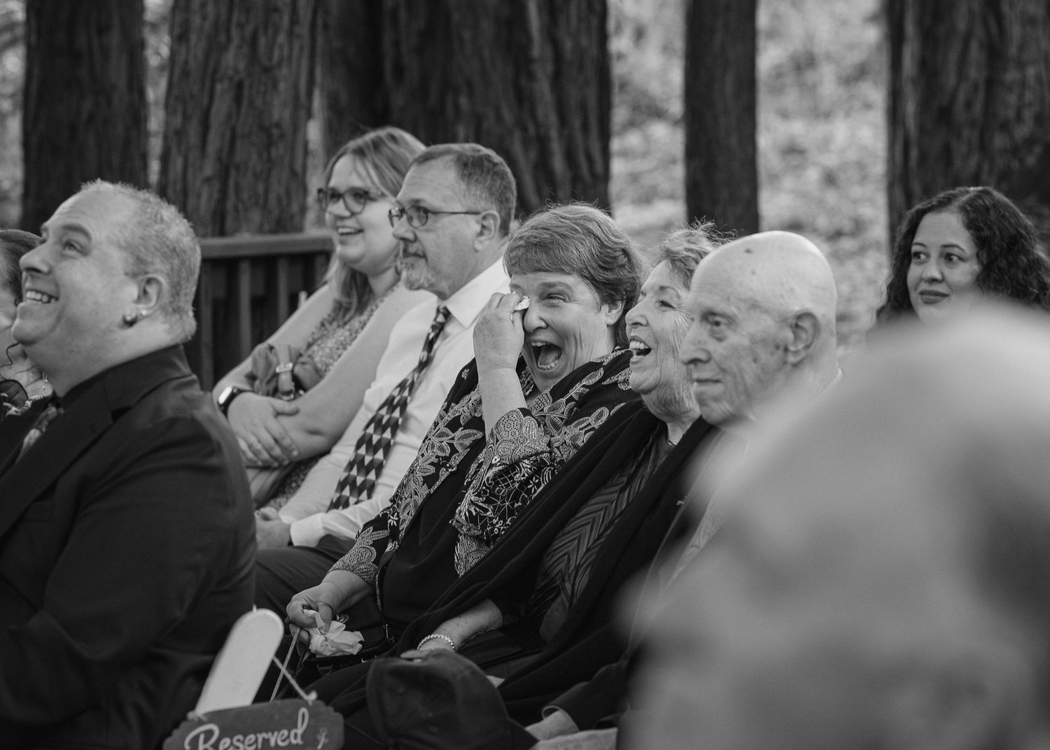 Black and white photo of smiling wedding guests seated outdoors among tall trees during a ceremony.