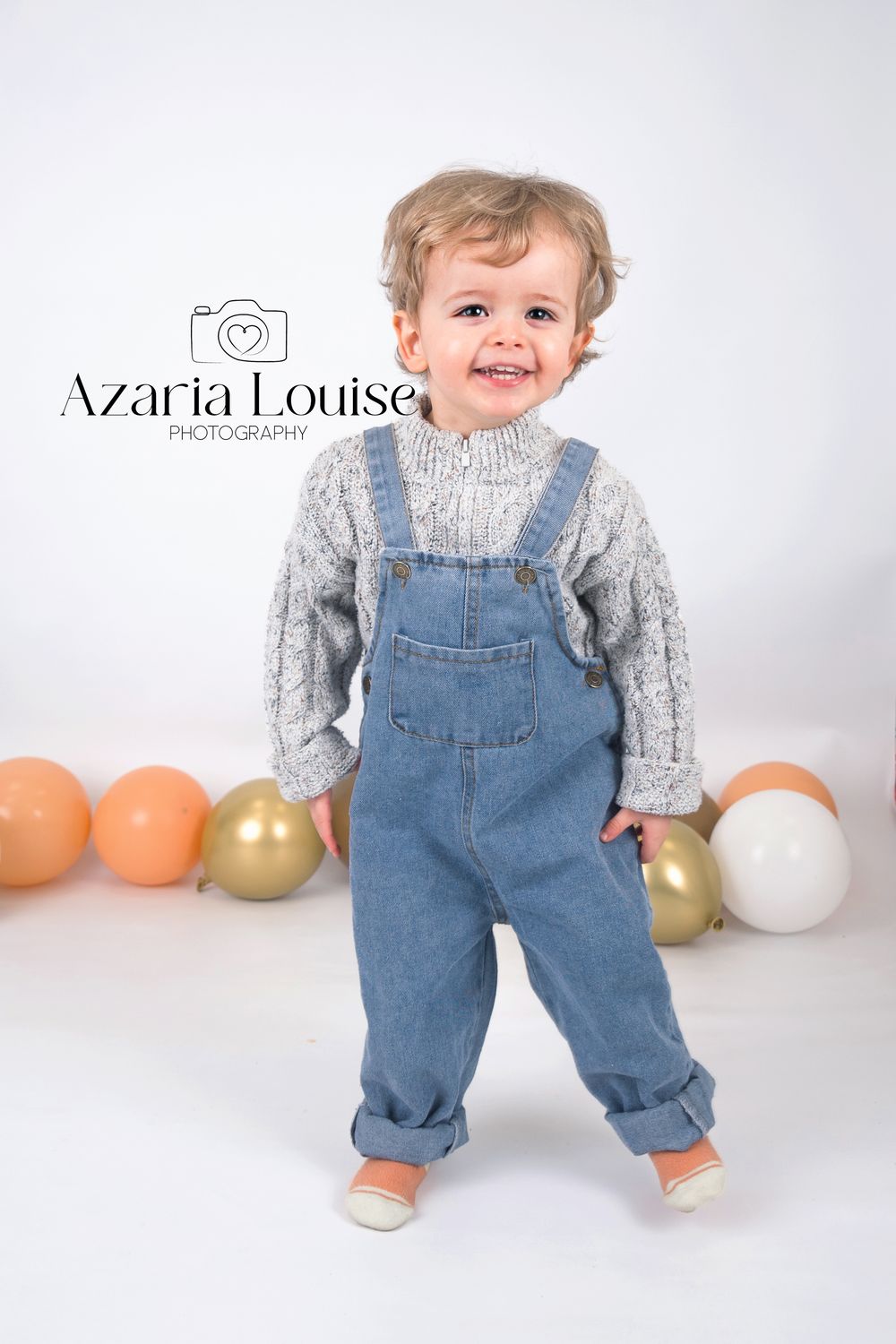 Toddler wearing gray sweater and denim overalls stands against white backdrop with orange and gold balloons.