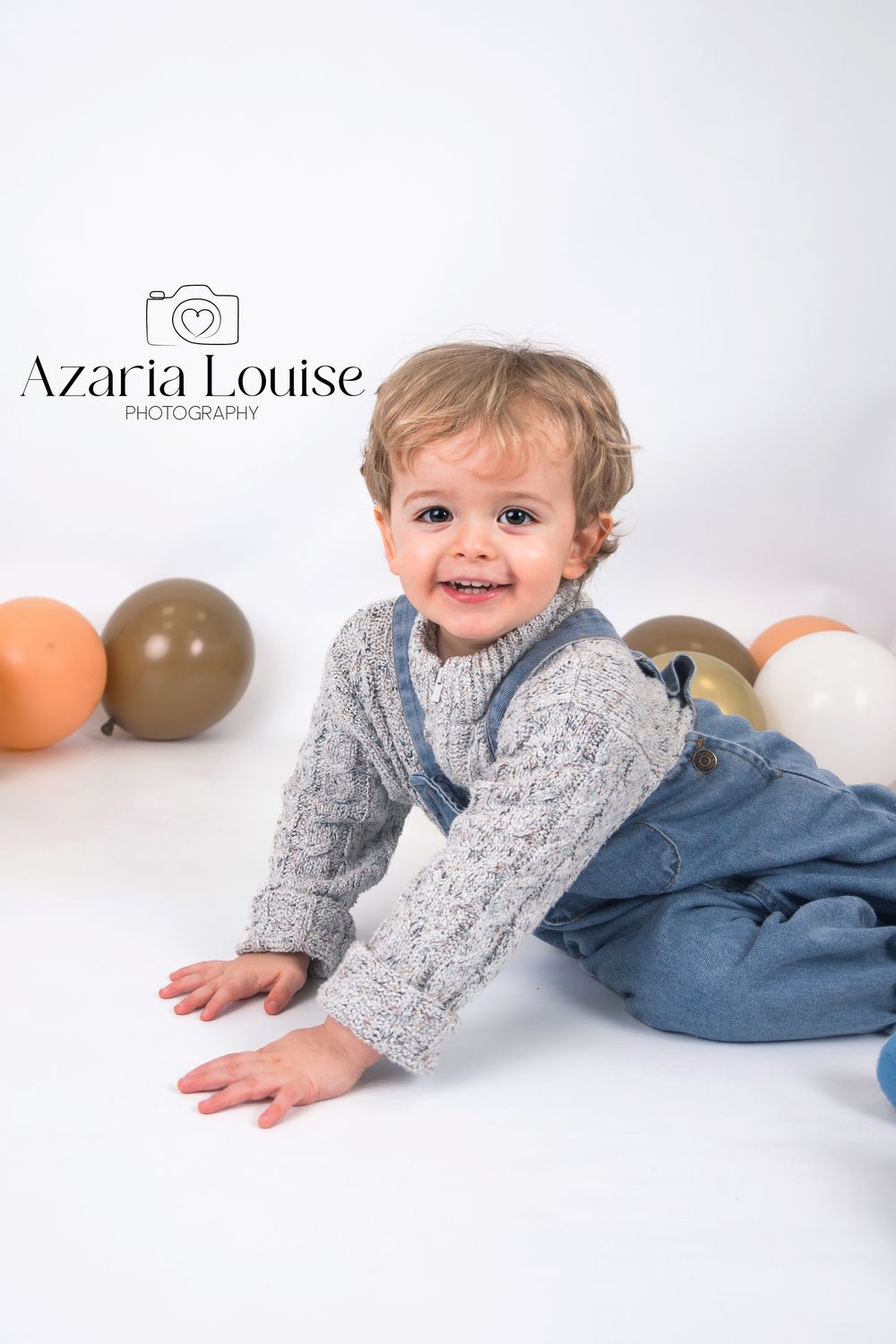 Smiling toddler in gray sweater and denim overalls crawls on white floor with colorful balloons in background.