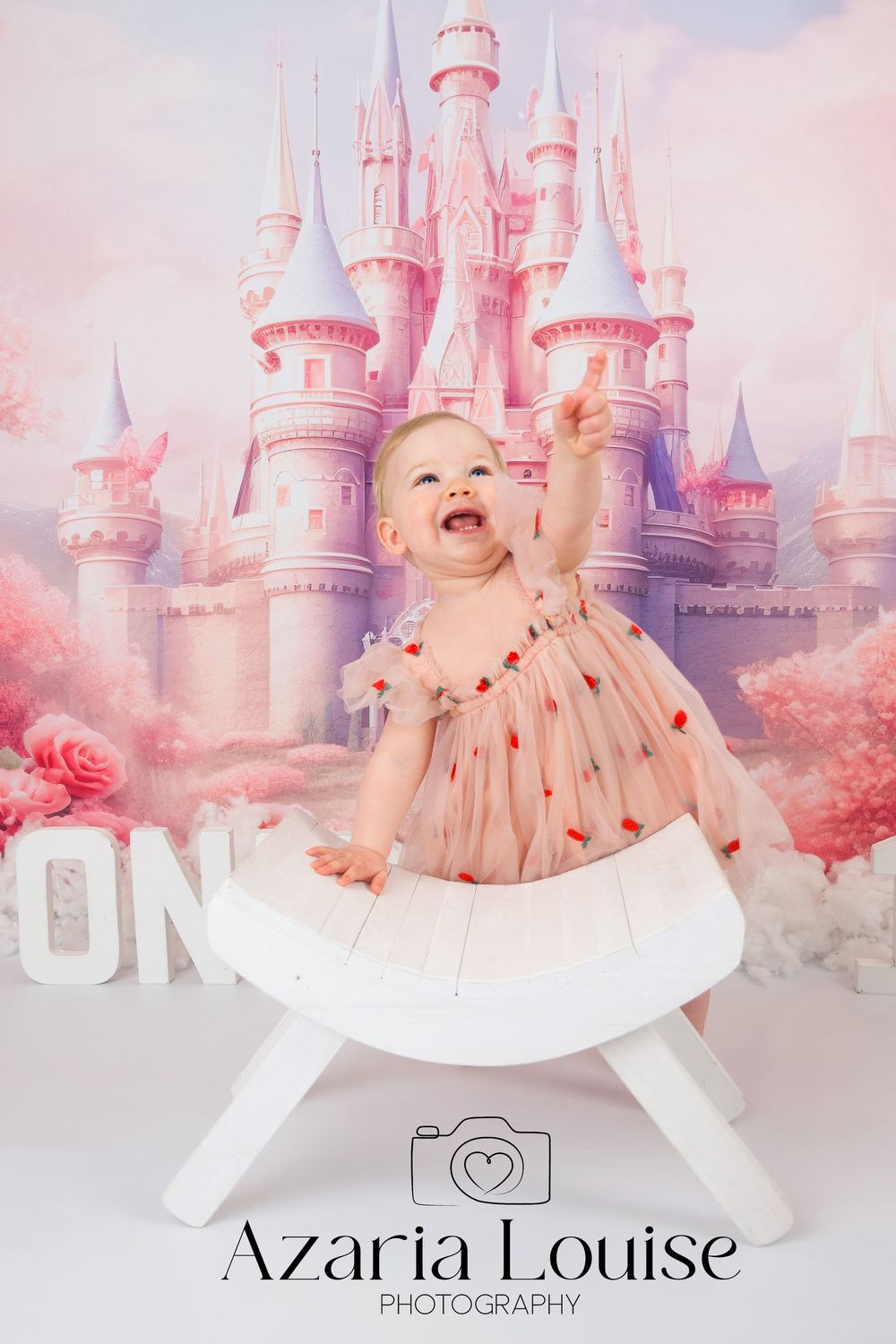 Baby in polka dot dress smiles with joy in front of pink fantasy castle backdrop during professional photoshoot.