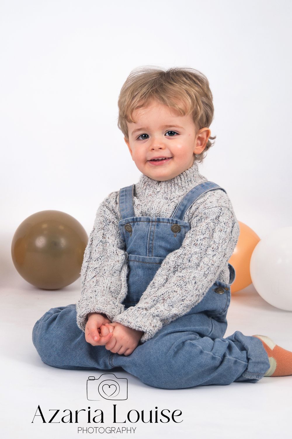 Child wearing jean overalls and gray sweater sits on floor with balloons during professional photo studio session.