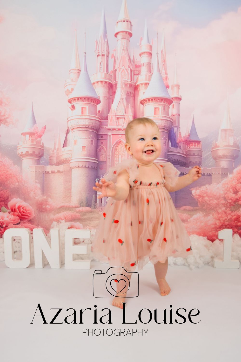 A toddler in a pink dress stands before a dreamy pink castle backdrop with roses and photography branding below.