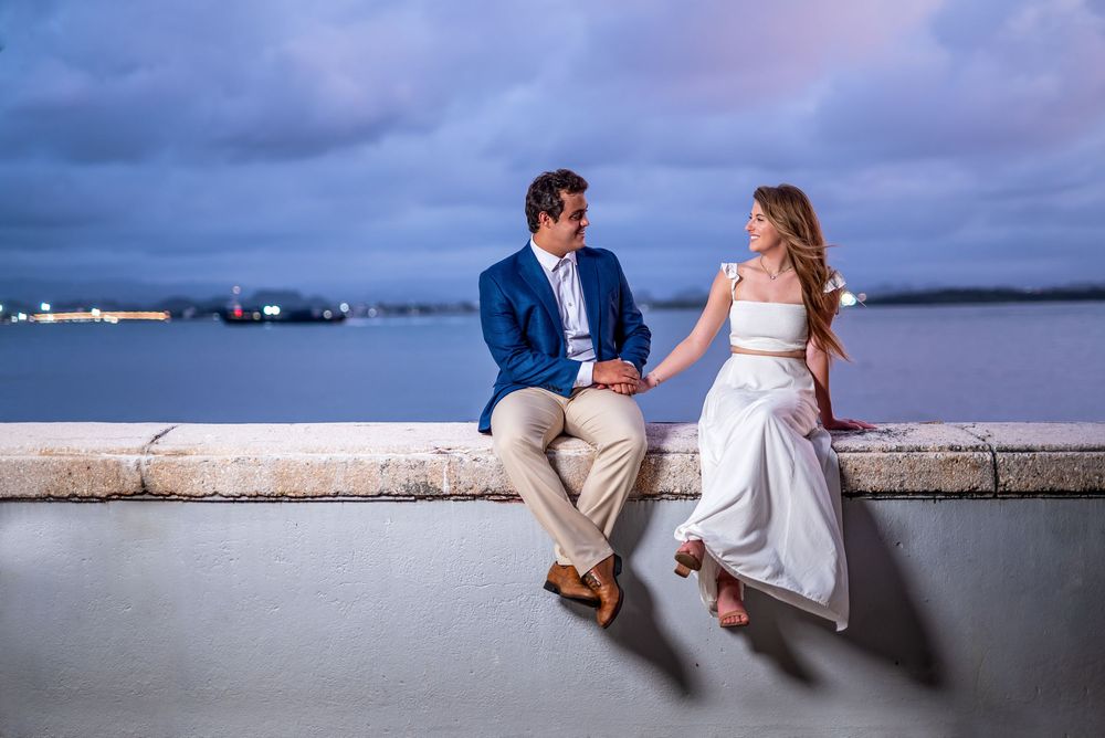 A couple sits on a waterfront wall at dusk, with a serene harbor view and moody purple clouds in the background.