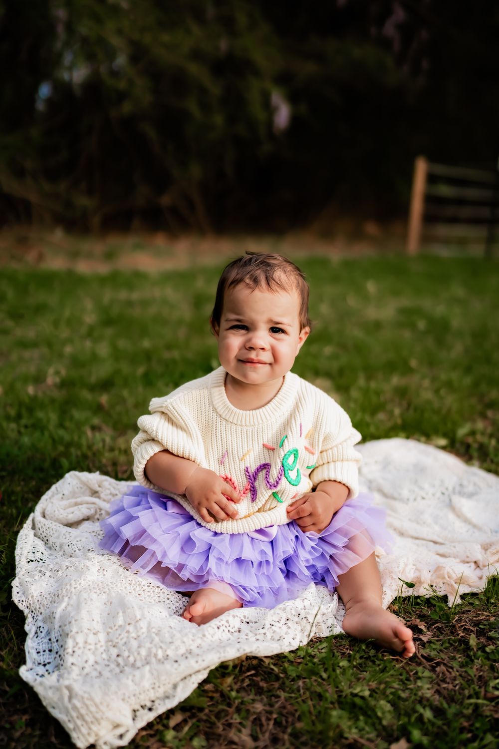 A small child in a cream sweater and purple tutu sits on a white lace blanket in the grass during a photo session.
