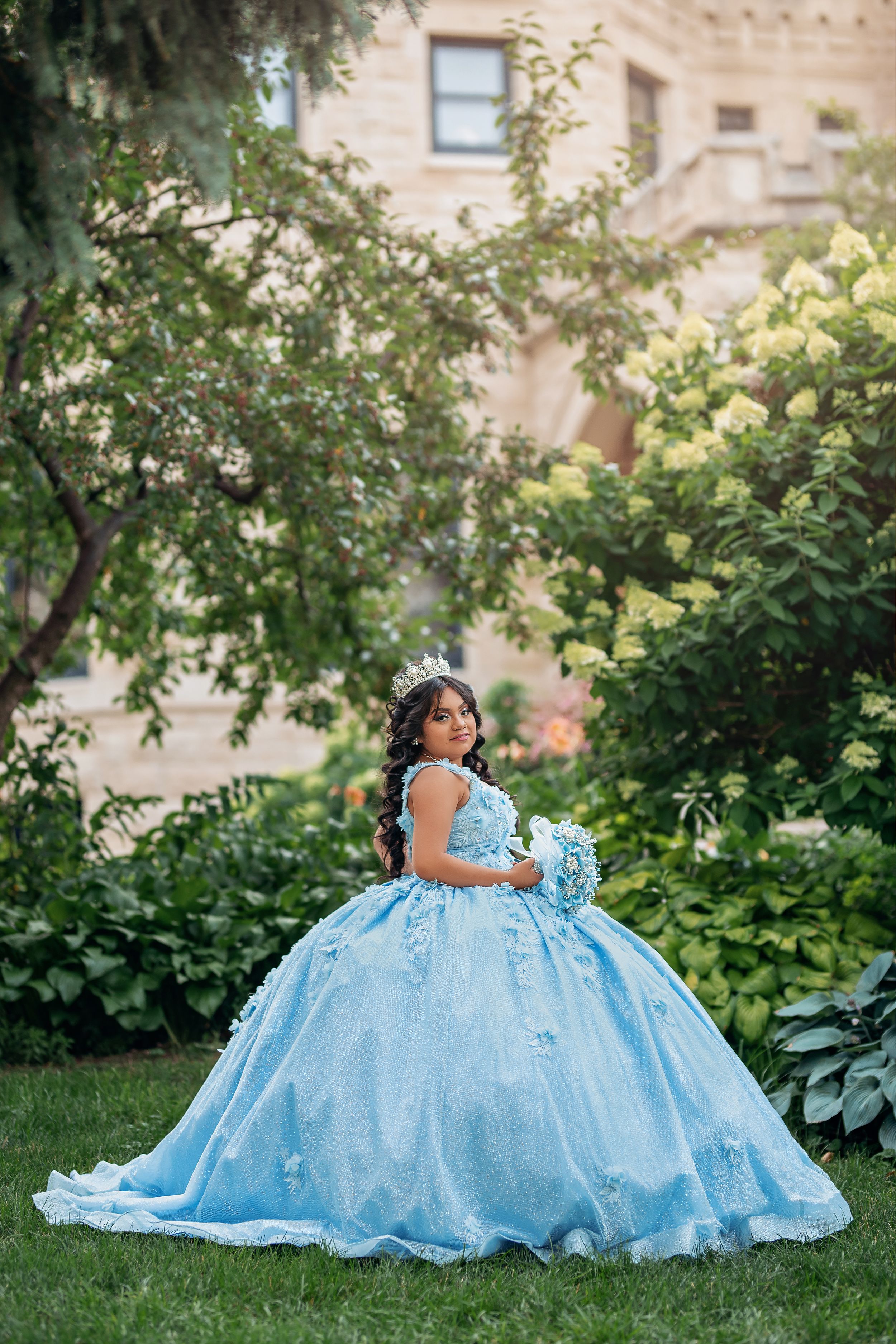 Light blue princess ballgown photographed in a lush garden setting with stone architecture.
