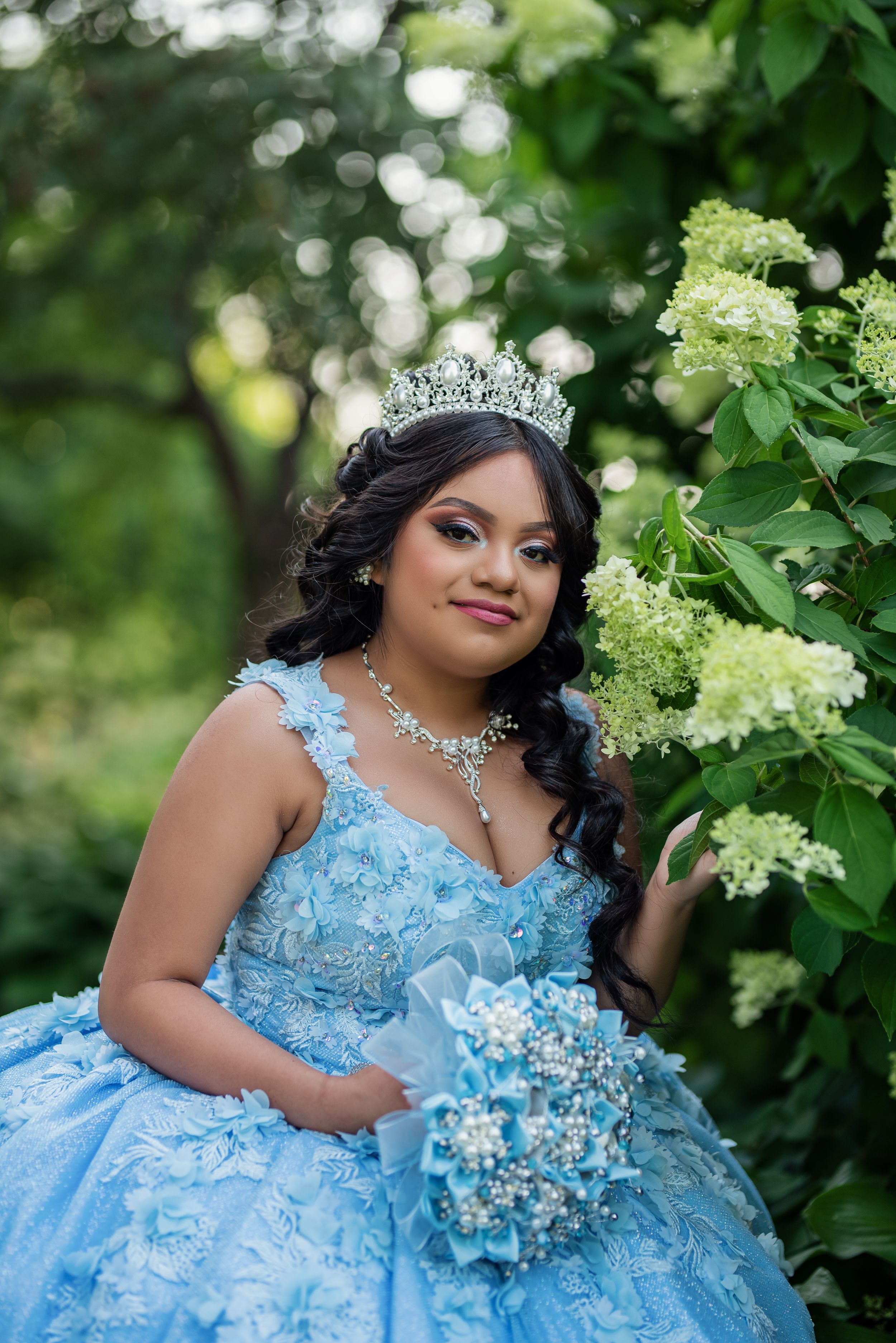 Powder blue quinceañera dress with detailed embellishments photographed among flowering bushes.