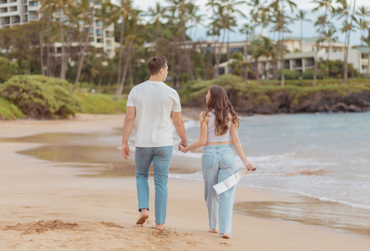 A couple embraces on a serene beach at sunset, with waves gently lapping at their feet and mountains in the background.