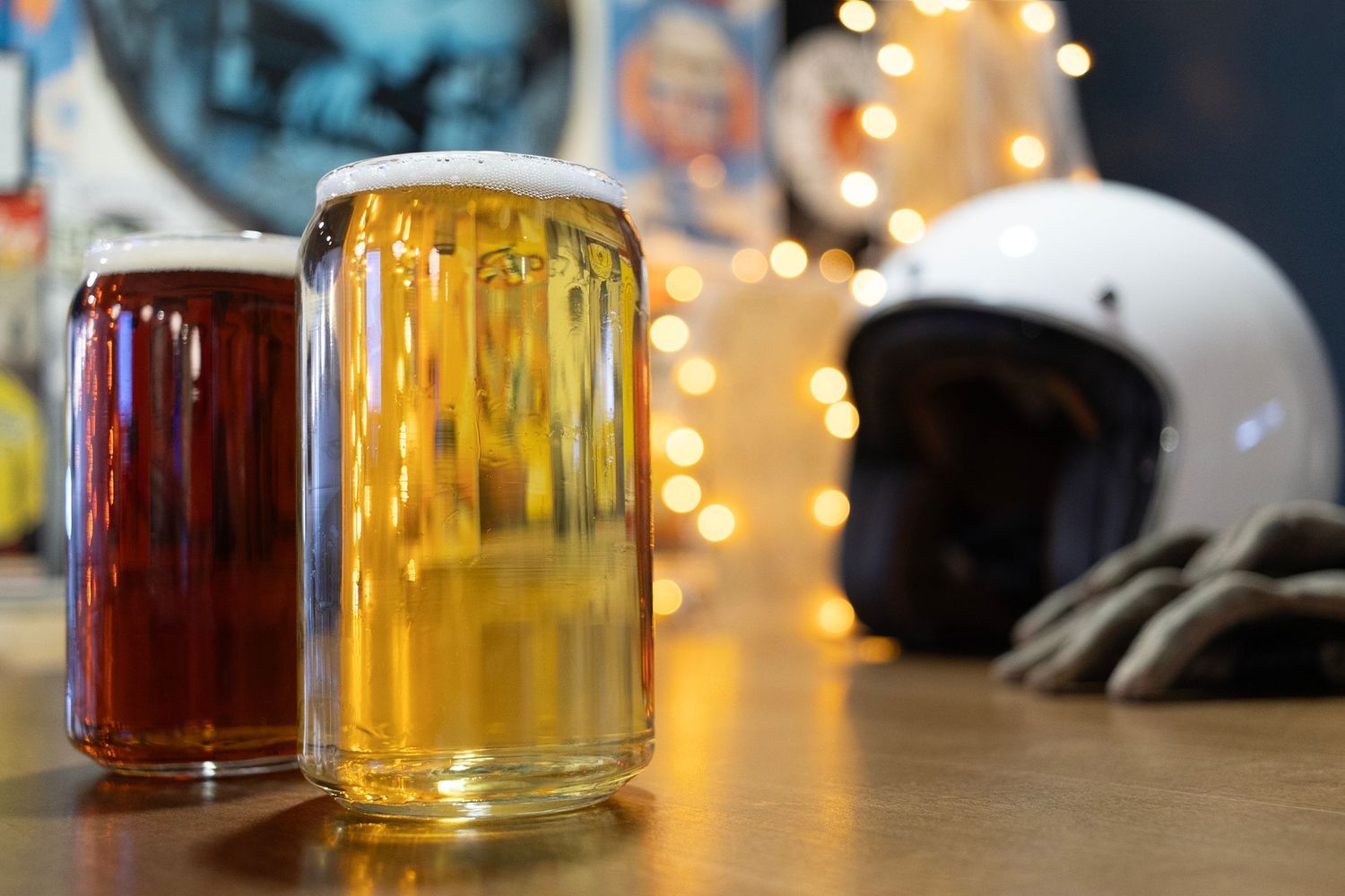Two craft beers in glasses with motorcycle helmet and string lights in background.