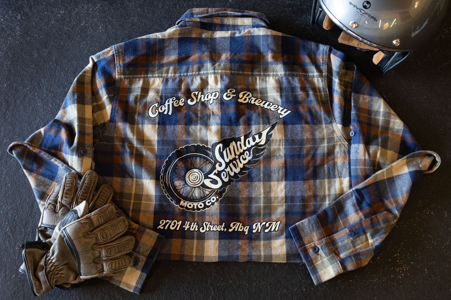 Plaid flannel shirt with Sunday branding and motorcycle helmet on dark surface.