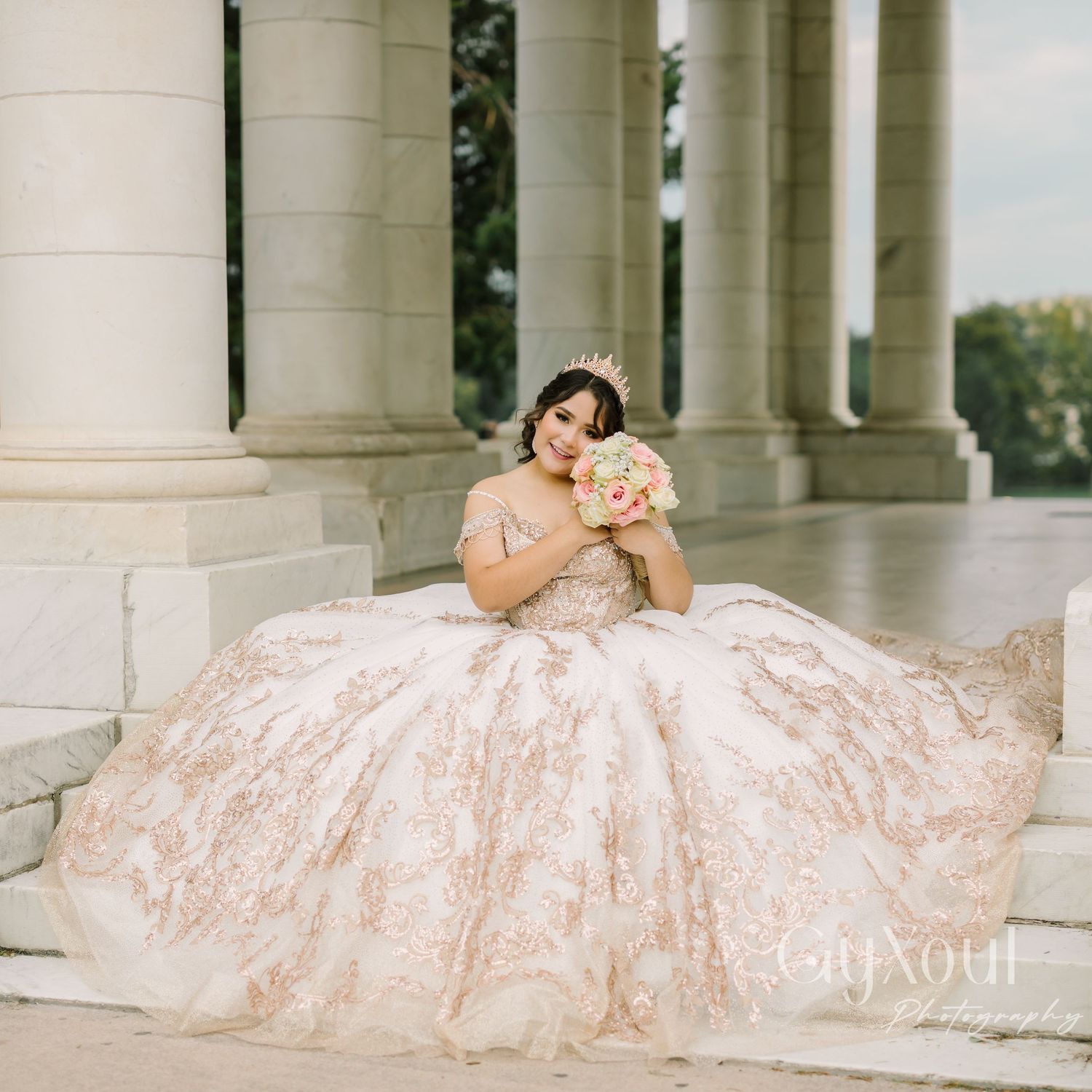 A bride in an ornate blush-colored ballgown sits gracefully against classic stone columns at an elegant venue.