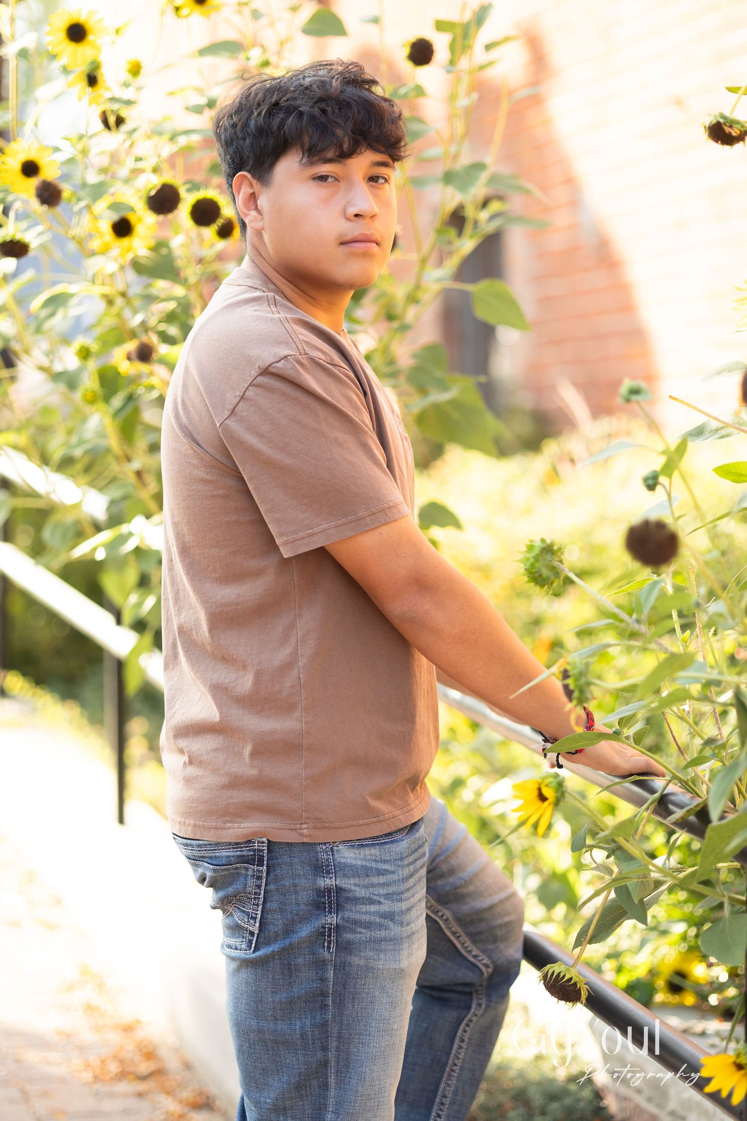 A person wearing a brown t-shirt and jeans poses in a garden with blooming sunflowers on a sunny afternoon.