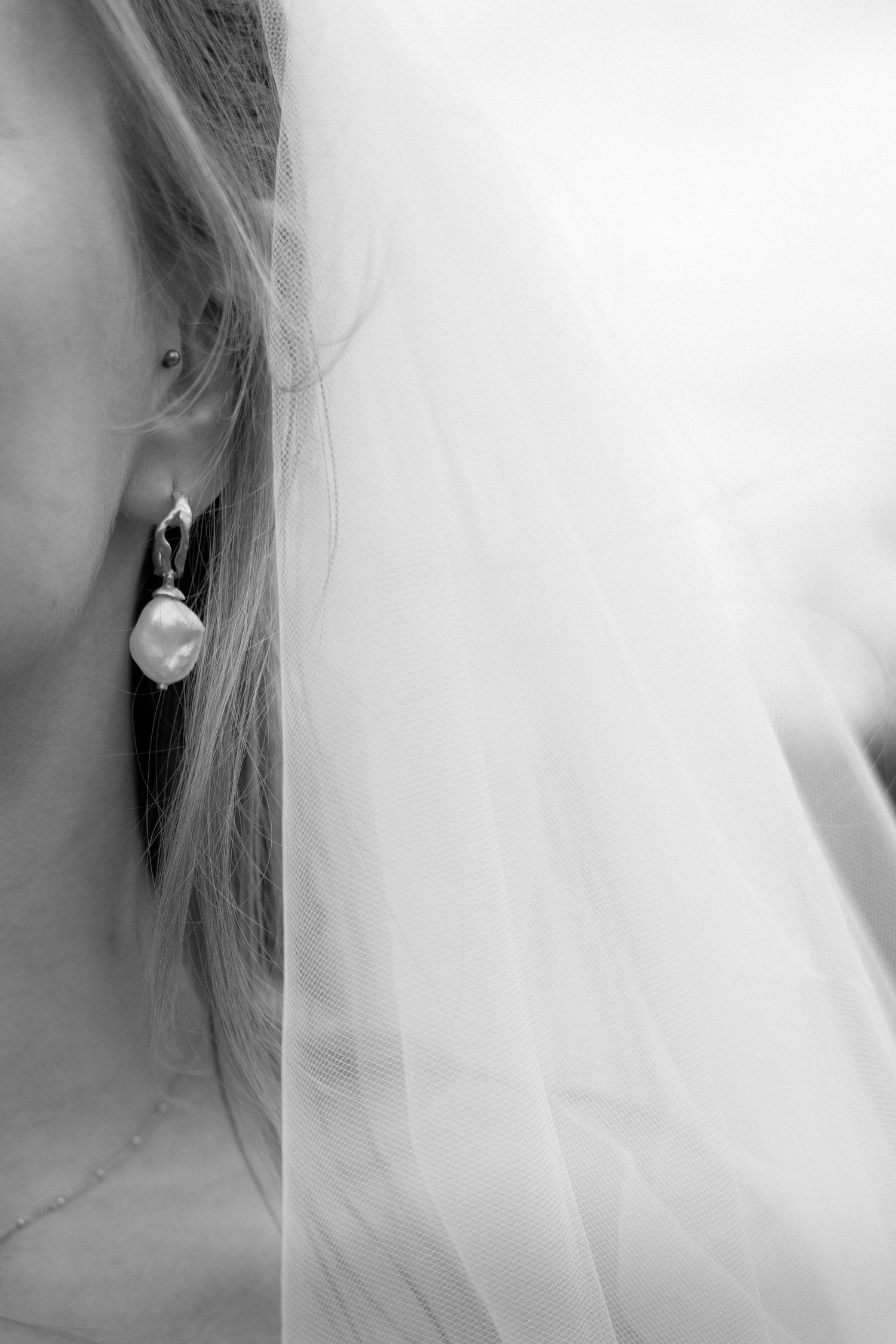 Black and white close-up of an elegant pearl drop earring with wispy blonde hair and delicate white veil in soft focus.