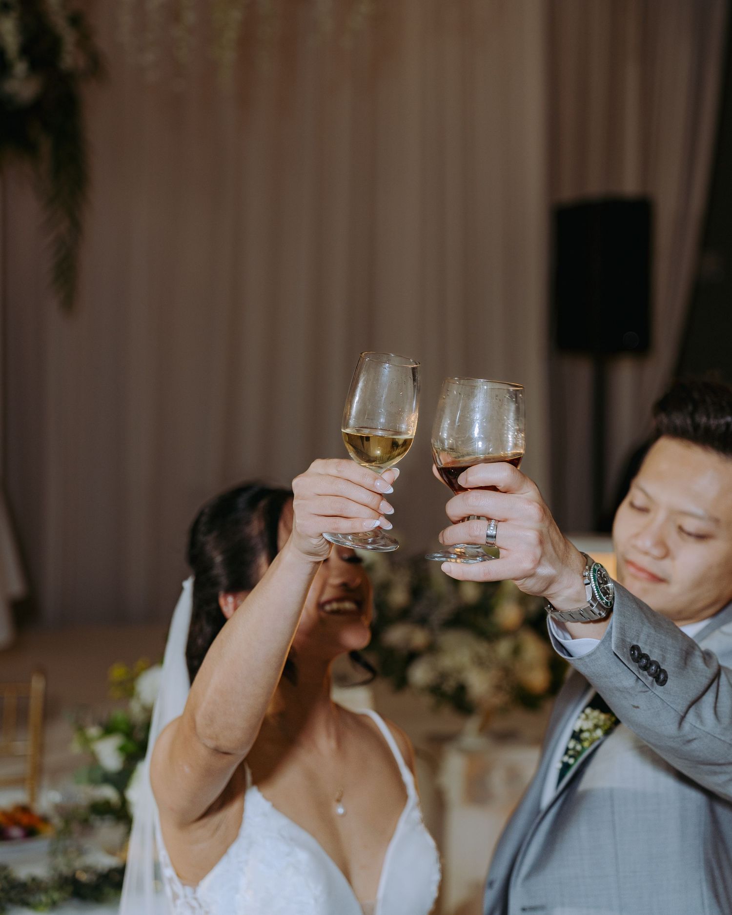 Newlyweds raise champagne glasses in celebration at wedding reception with soft lighting and floral decor.