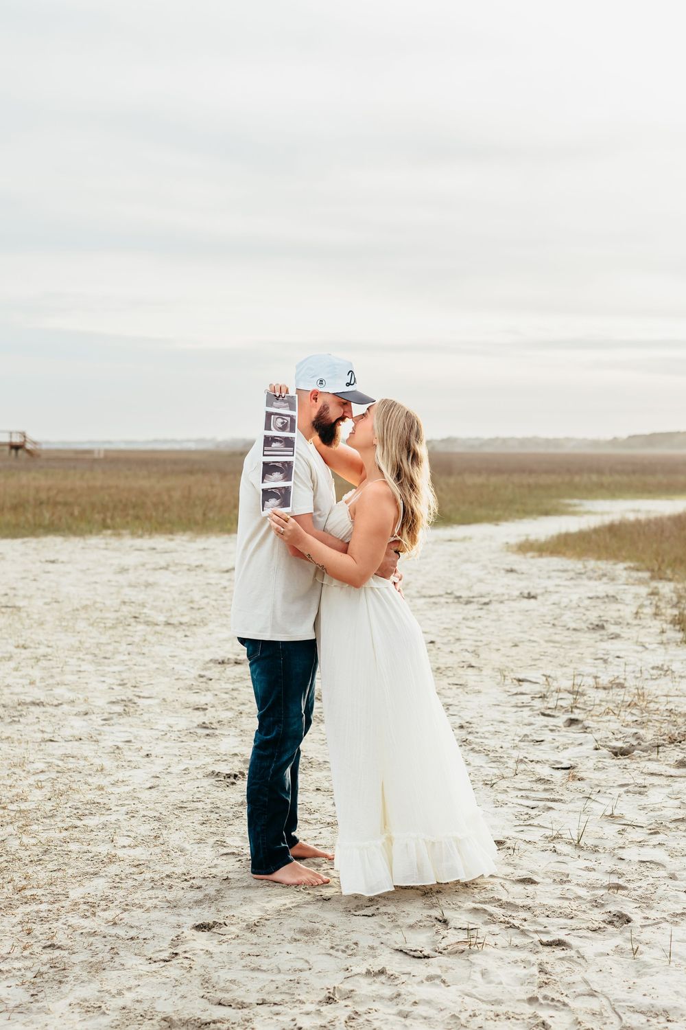 Couple sharing romantic moment while holding ultrasound photos on coastal beach.