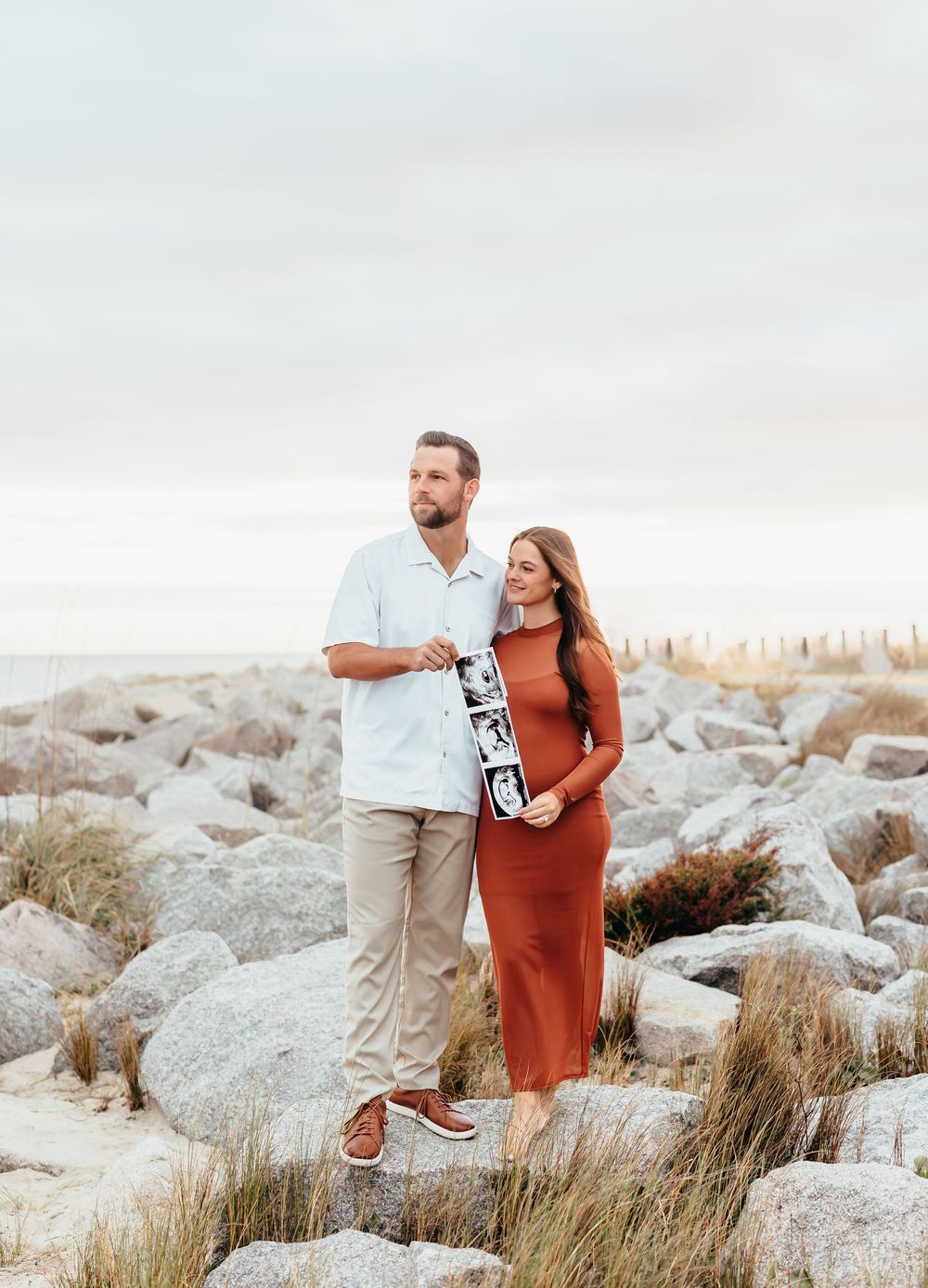 Couple in white and rust colored outfits standing among coastal rocks with sonogram photos.