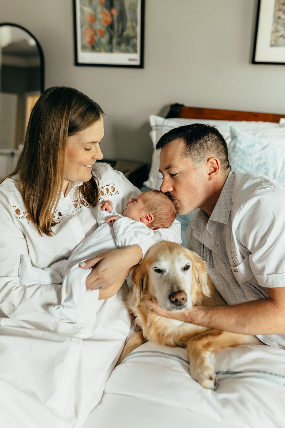 A couple sits on a bed with their newborn baby and golden retriever dog, the father kissing the infant tenderly.