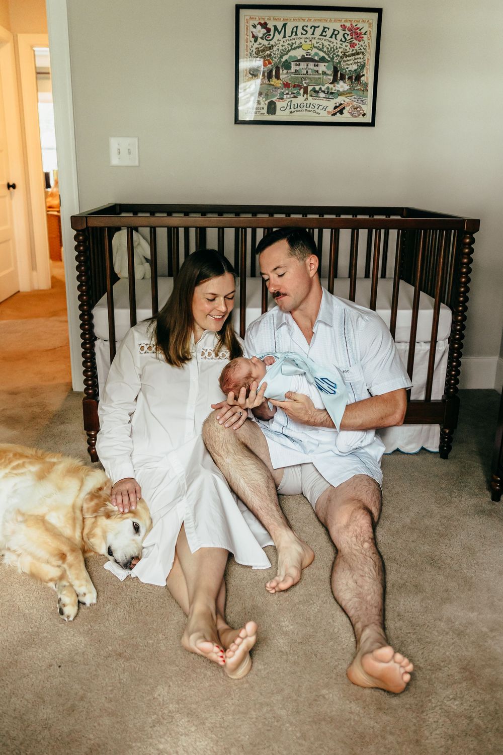 New parents sit on nursery floor with newborn baby and golden retriever dog beside a dark wood crib.