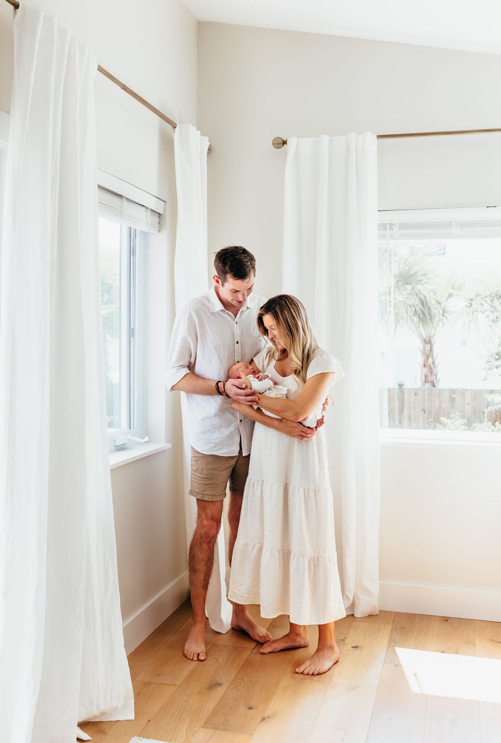 A couple stands in a bright room with white curtains, holding their newborn baby.