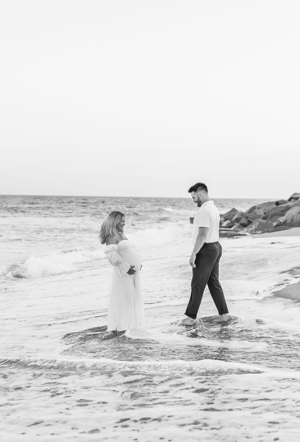Couple standing in shallow ocean waves during black and white beach photoshoot.