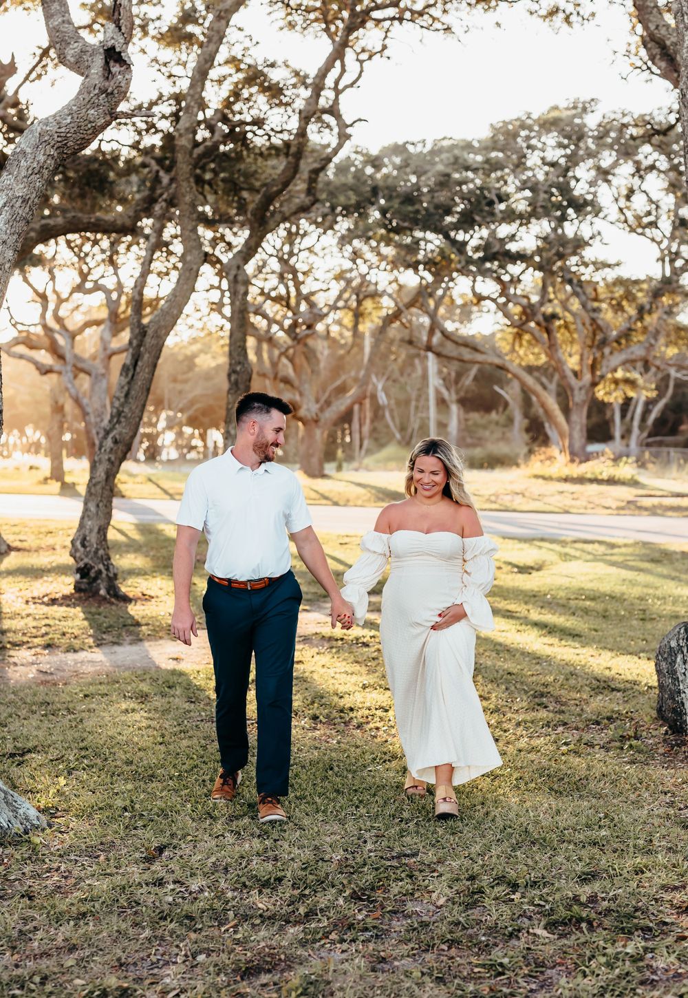 A couple strolls hand in hand through a park with sun-dappled trees in the background.