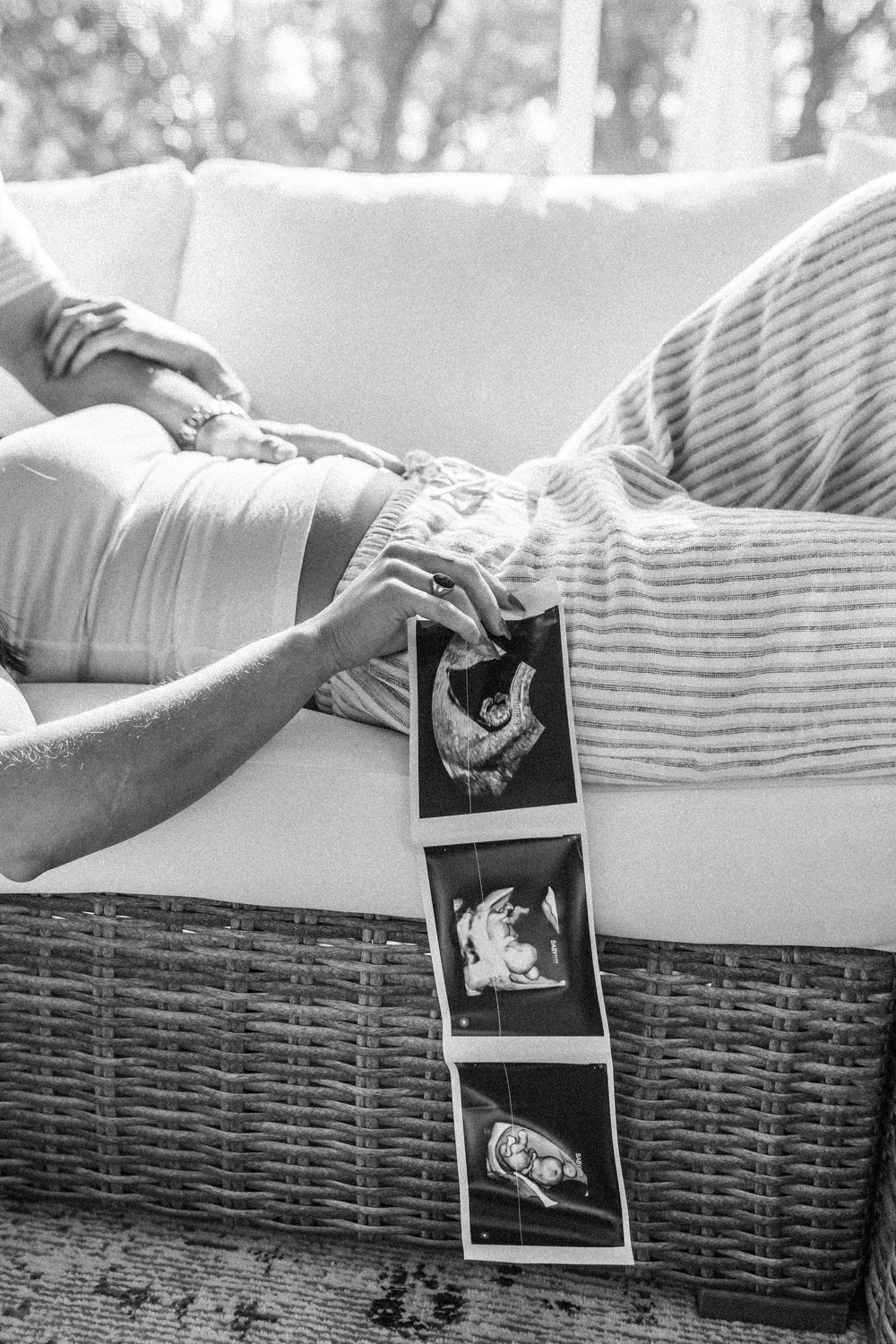 Black and white photo of pregnant woman holding ultrasound images while sitting on a wicker sofa outdoors.