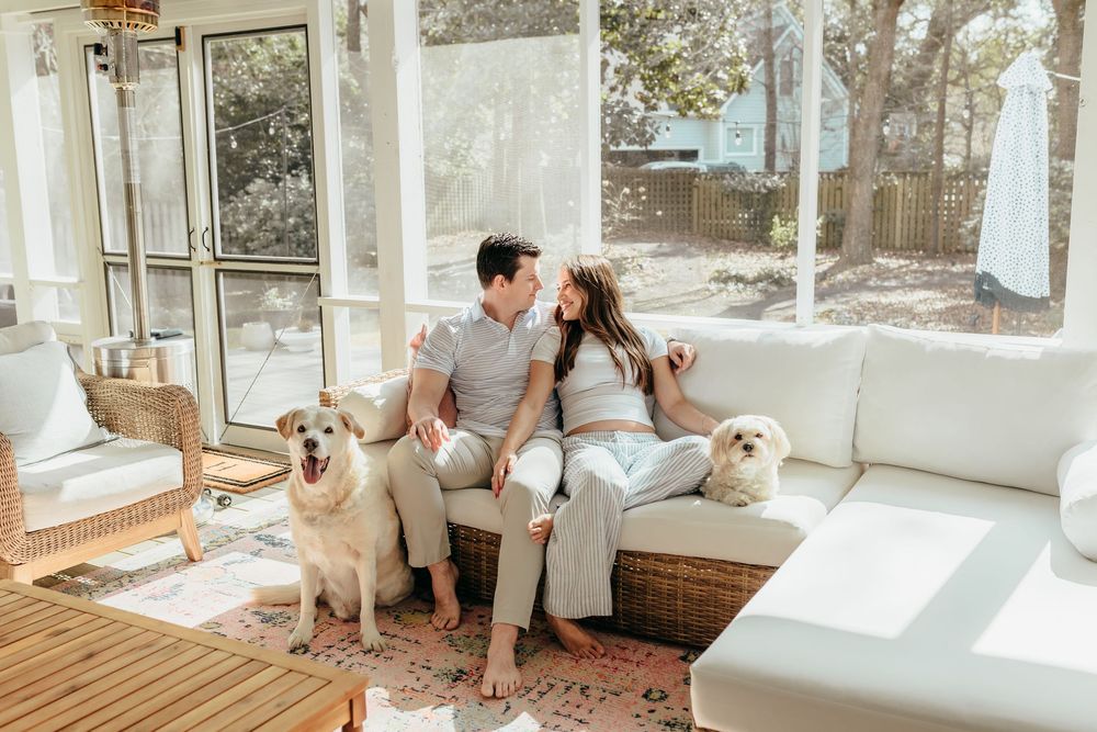 Couple sitting together on white couch in sunlit sunroom with two golden dogs nearby.