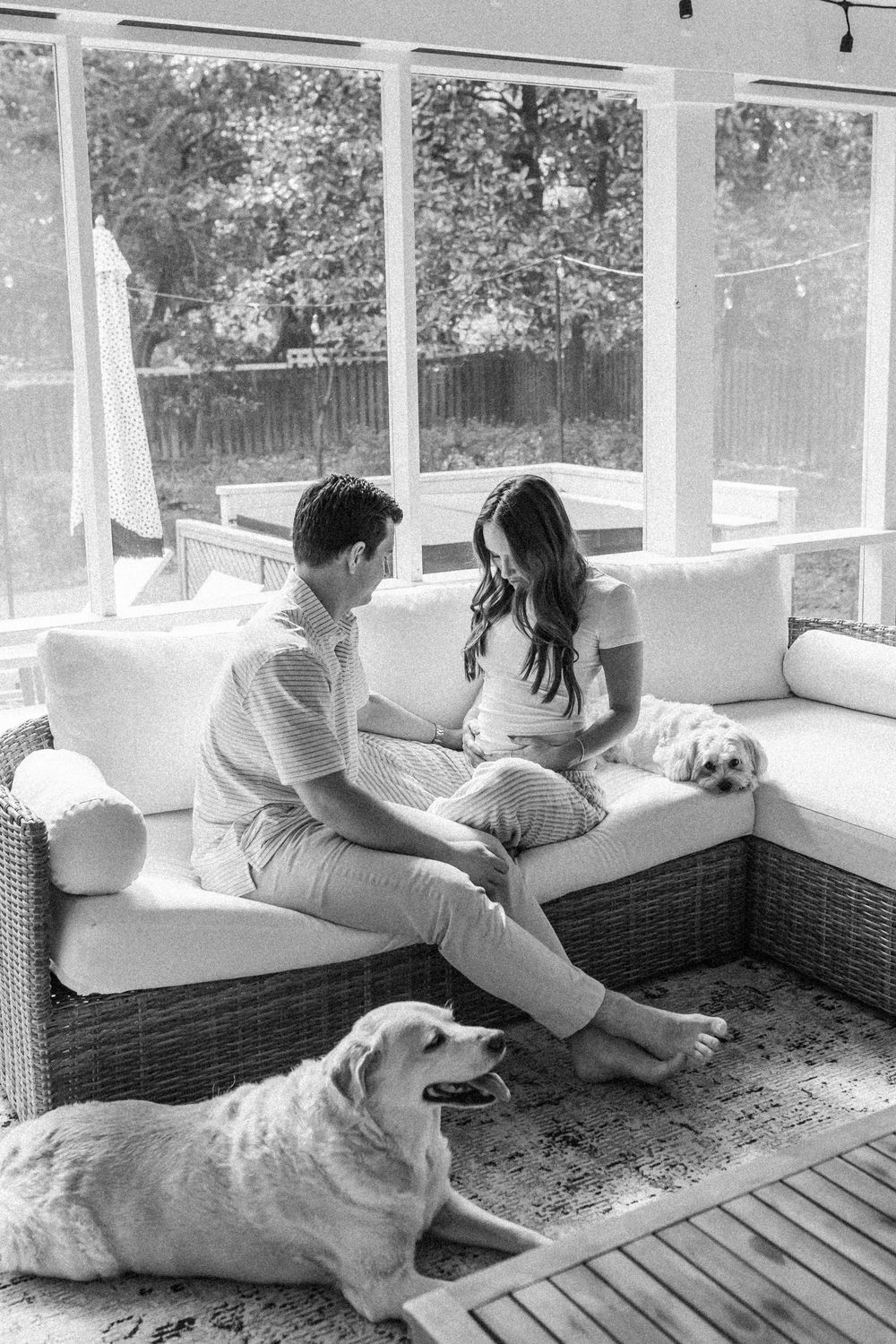 Black and white photo of a couple sitting on a wicker porch sofa with two dogs, one resting on the floor.