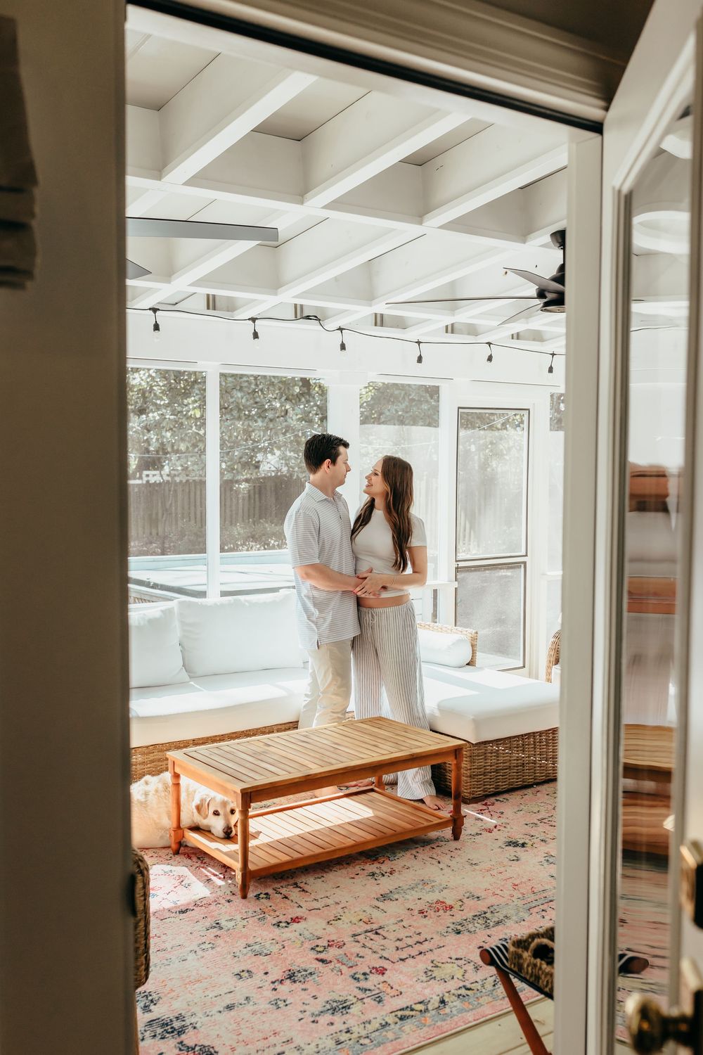 A couple embraces in a bright sunroom with exposed beams, string lights, and a cozy seating area on a floral rug.