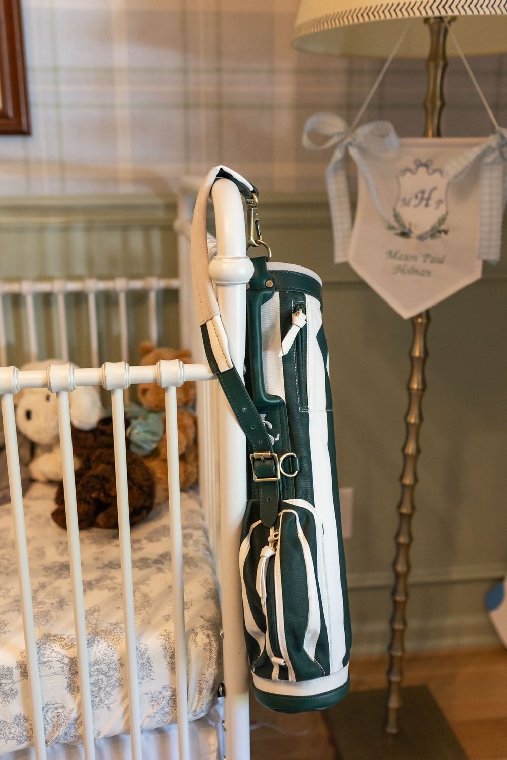 Green and white golf bag leaning against a white baby crib in a plaid nursery room with a personalized pennant.