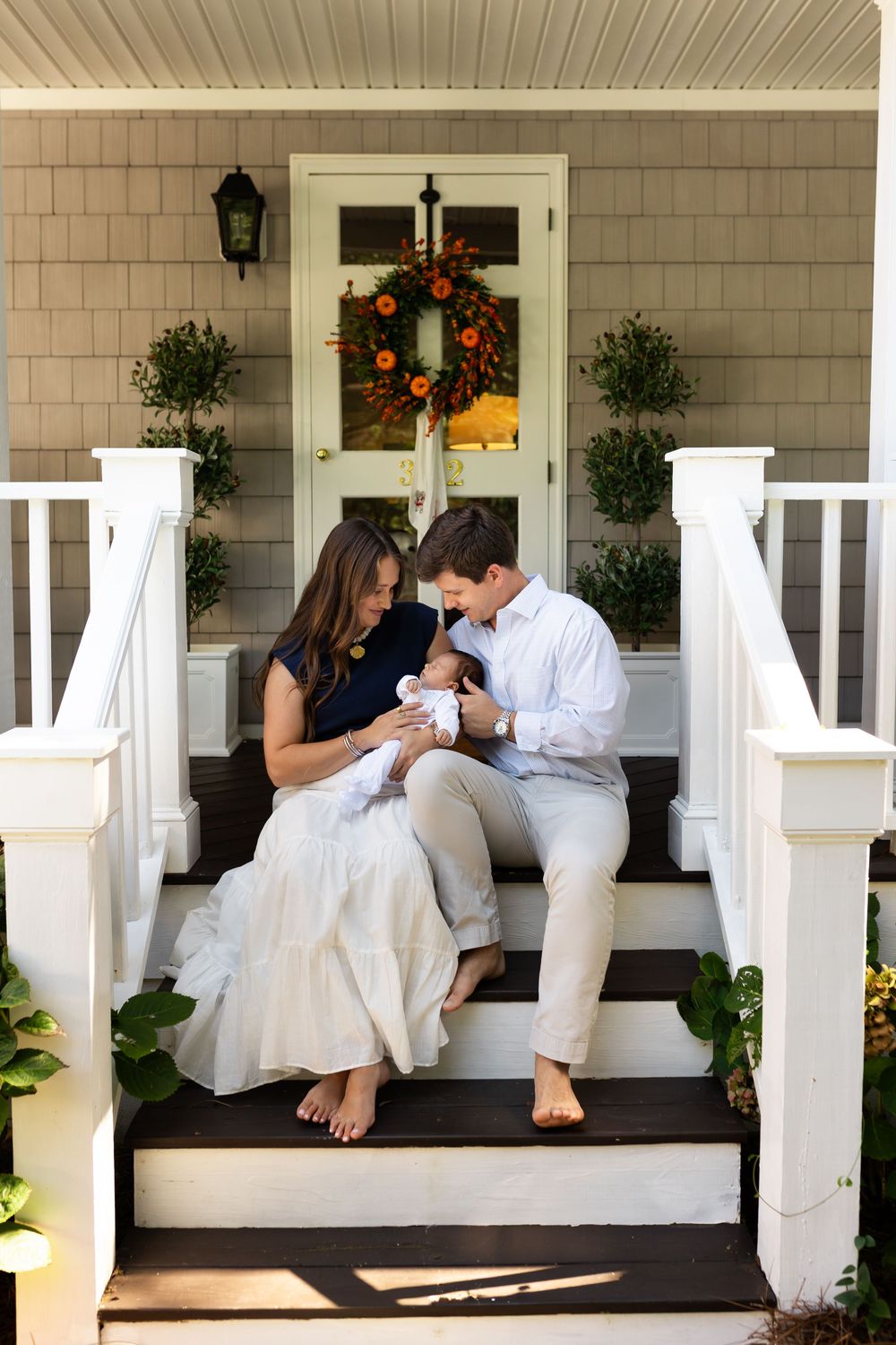 Young couple sitting on front porch steps admiring their newborn baby, surrounded by fall decor.