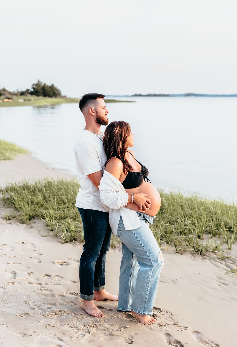 A romantic couple embraces on a scenic beachfront with grass and water views.