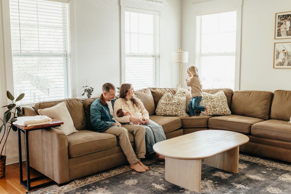 Family relaxing in bright living room on large brown sectional sofa with newborn baby and young child playing nearby.