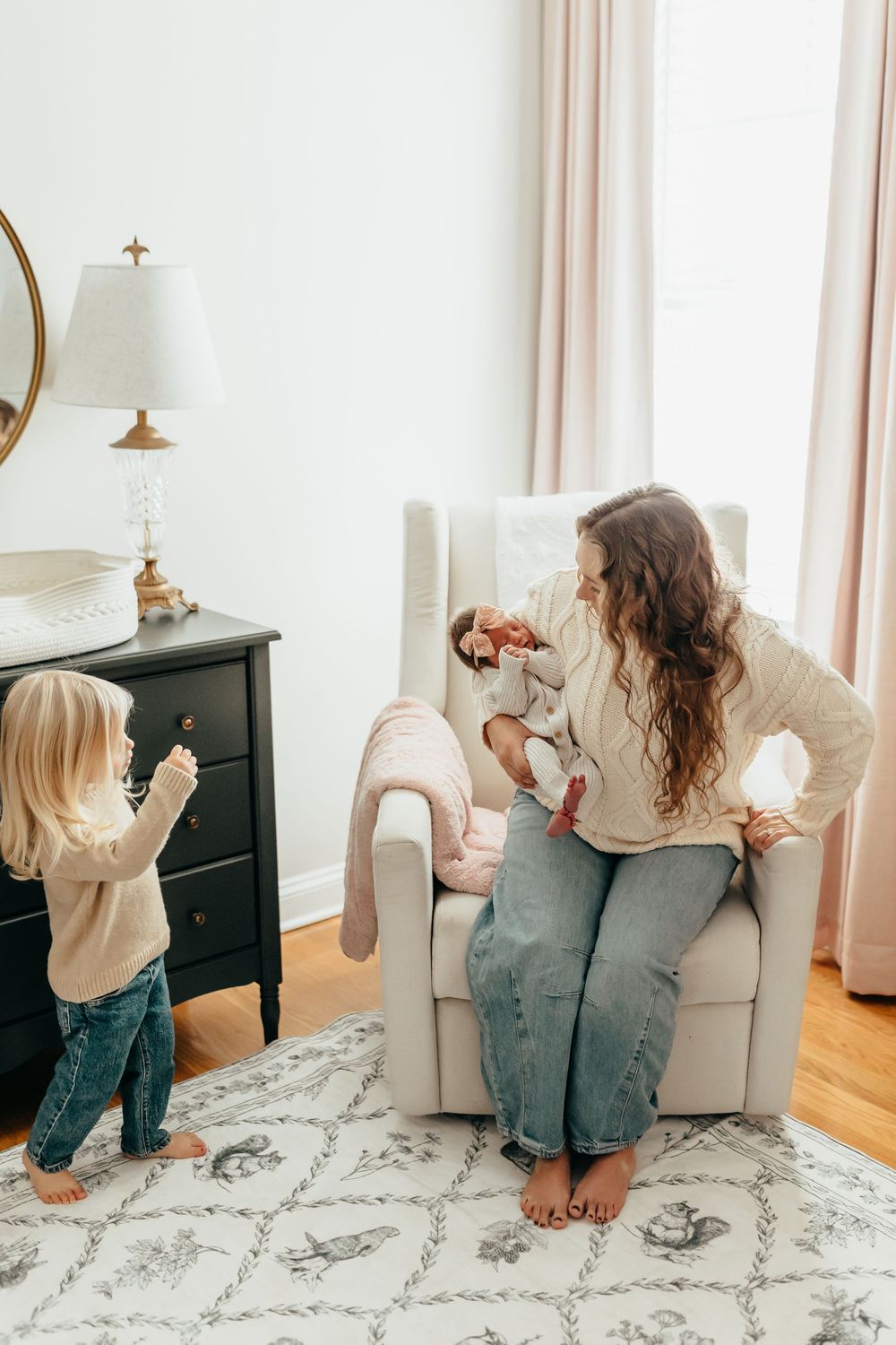 Mother sits in nursery glider holding newborn while toddler plays nearby on patterned rug in bright room.